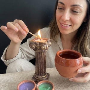 May include: A lit candle in a decorative, brown, column-shaped holder. Two small, terracotta-coloured bowls with purple and green wax candles are in front. A person is holding a terracotta-coloured cup.