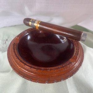 May include: A wooden ashtray with a dark brown interior and a decorative outer rim. A brown cigar with a gold band rests on the ashtray. The ashtray and cigar are set against a white background.