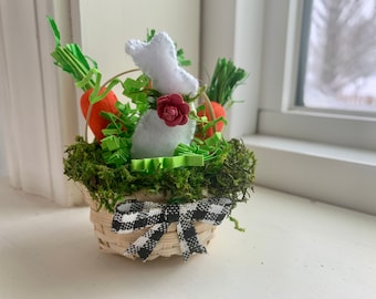 May include: A small white felt bunny sits in a woven basket with green grass, a red flower, and two orange carrots. The basket has a black and white checkered ribbon.