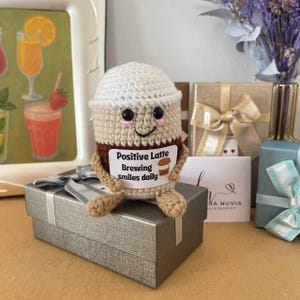 Könnte beinhalten: Gehäkelter Latte-Becher mit lächelndem Gesicht und dem Text "Positive Latte Brewing smiles daily" auf einem Etikett. Der Becher sitzt auf einer silbernen Geschenkbox. Im Hintergrund befinden sich weitere Geschenkboxen und ein Tablett mit Getränkeillustrationen.