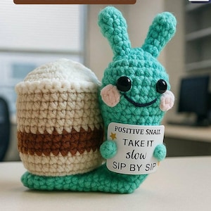 May include: A crocheted snail with a teal body, cream shell, and brown accents. The snail holds a sign that reads "POSITIVE SNAIL TAKE IT slow SIP BY SIP." The image also includes the text "Crochet Pattern".