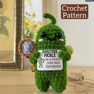 Puede incluir: Una figura de pepinillo verde tejido a ganchillo sosteniendo una raqueta de tenis y un letrero que dice "POSITIVE PICKLE IN A PICKLE? JUST KEEP SWINGING!". La imagen también incluye el texto "Crochet Pattern" y un icono PDF.
