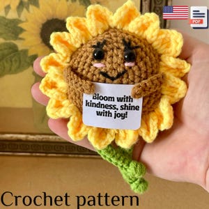 May include: A crocheted sunflower with yellow petals, a brown center, and a green stem. The sunflower holds a white sign with the text "Bloom with kindness, shine with joy!" The crochet pattern is available.