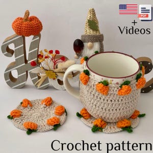 May include: A crochet pattern for a fall-themed mug cozy and matching coasters. The cozy and coasters are beige with orange pumpkin accents. The image also includes a decorative wooden letter, a gnome figurine, and the text "Crochet pattern".