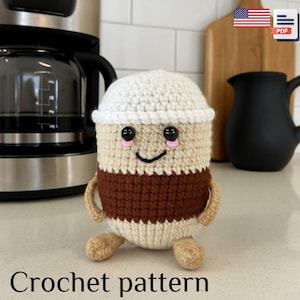 May include: A crocheted coffee cup toy with a white top, a tan body, and a brown band. It has black eyes, pink cheeks, and a smiling mouth. The arms and feet are tan. The text "Crochet pattern" is at the bottom.
