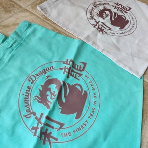 May include: Two tote bags with a teal and white background. Both bags feature a brown logo with a dragon and the text "Jasmine Dragon The Finest Teas in Ba Sing Se".