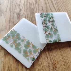 May include: Two square glass coasters with a white background and a green and gold leaf design. The design is on one side of each coaster.