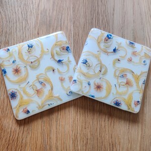 May include: Two square white coasters with a gold and blue floral pattern. The coasters have a white background with gold swirls and blue flowers.