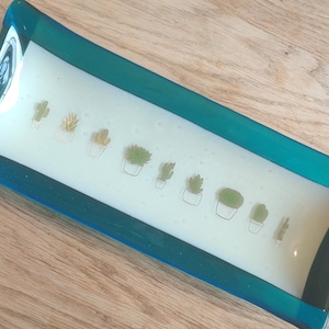 May include: A teal glass dish with a white interior featuring a line of potted succulents. The succulents are drawn in gold and green.