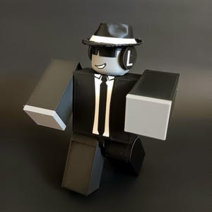 May include: A blocky, articulated figure with a gray head, wearing a black fedora with a white band, black sunglasses, and black headphones with a white letter L. The figure has a black suit, white shirt, and black tie, with gray hands and legs.