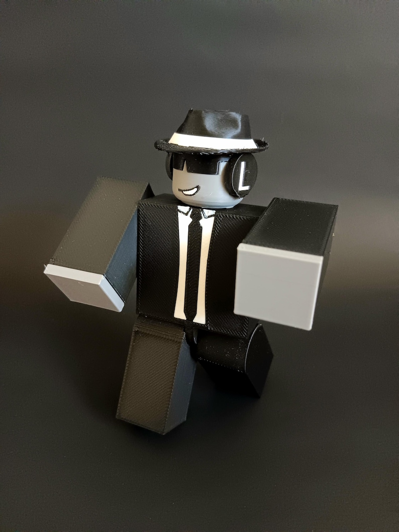 May include: A blocky, articulated figure with a gray head, wearing a black fedora with a white band, black sunglasses, and black headphones with a white letter L. The figure has a black suit, white shirt, and black tie, with gray hands and legs.