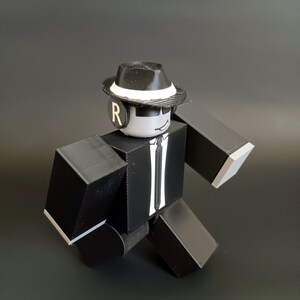 May include: A blocky, articulated action figure wearing a black suit with a white tie, a black fedora with a white band, and black sunglasses. The figure has a white head with a smiling expression and a black circular earpiece featuring the letter R.