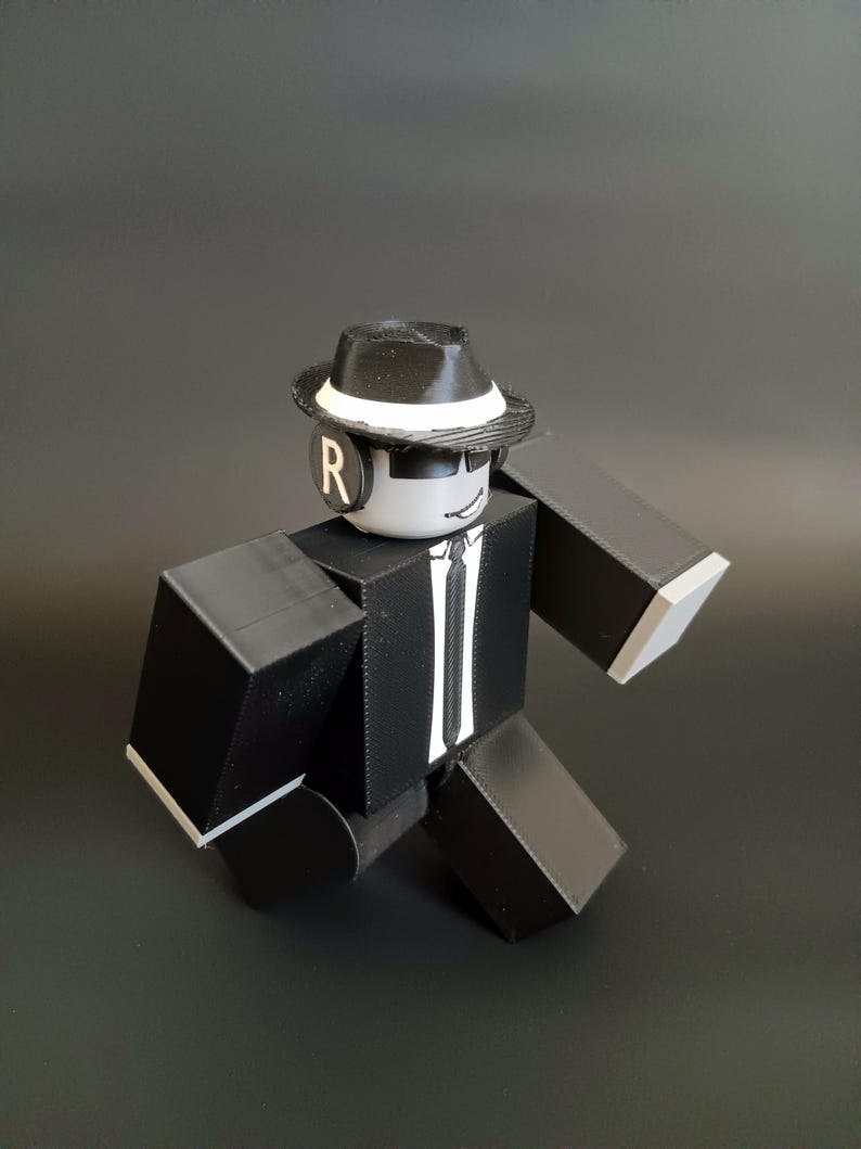 May include: A blocky, articulated action figure wearing a black suit with a white tie, a black fedora with a white band, and black sunglasses. The figure has a white head with a smiling expression and a black circular earpiece featuring the letter R.