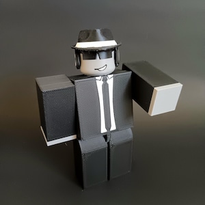 May include: A blocky, three-dimensional figure wearing a black suit, a white shirt with a black tie, and a black fedora with a white band. The figure has a gray head with black sunglasses and a simple white mouth line, featuring a textured surface.