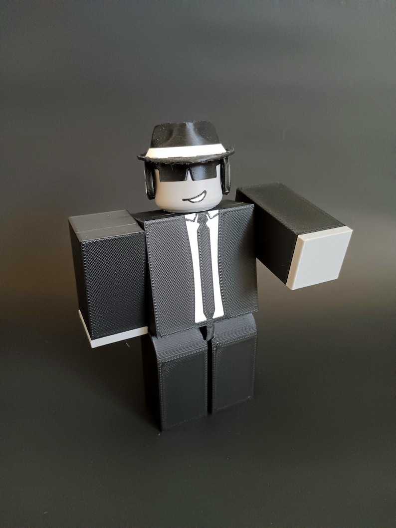 May include: A blocky, three-dimensional figure wearing a black suit, a white shirt with a black tie, and a black fedora with a white band. The figure has a gray head with black sunglasses and a simple white mouth line, featuring a textured surface.