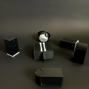 May include: A collection of black 3D printed plastic components including four rectangular blocks and one central piece featuring a white tie graphic, accompanied by a small white circular head figure wearing a black hat with a white letter L.
