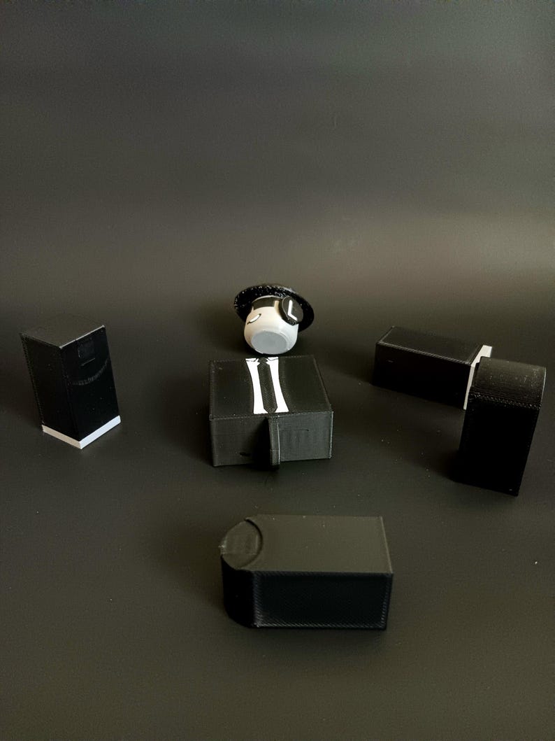 May include: A collection of black 3D printed plastic components including four rectangular blocks and one central piece featuring a white tie graphic, accompanied by a small white circular head figure wearing a black hat with a white letter L.