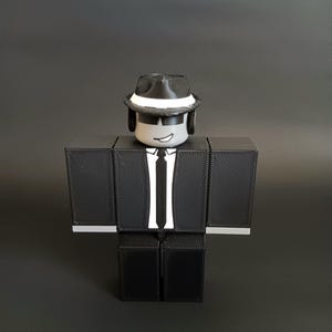 May include: A blocky, three-dimensional figure with a gray head, black sunglasses, and a black fedora with a white band. The figure wears a black suit jacket with a white shirt, a black tie, and black pants, featuring a printed smirk on the face.