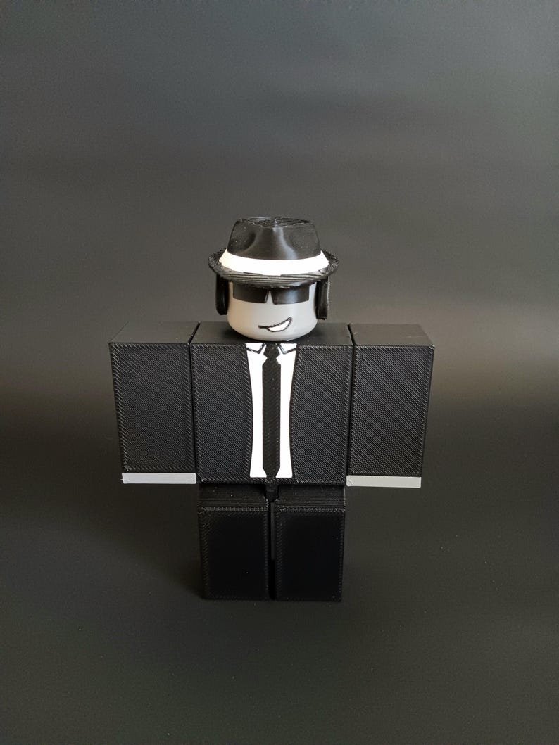 May include: A blocky, three-dimensional figure with a gray head, black sunglasses, and a black fedora with a white band. The figure wears a black suit jacket with a white shirt, a black tie, and black pants, featuring a printed smirk on the face.