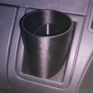 May include: A black, 3D-printed car cup holder. The cup holder is cylindrical with a slightly tapered design and fits into a car's existing console. The holder is designed to securely hold beverages while driving.