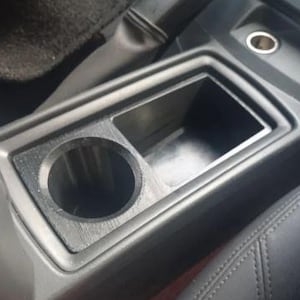 Könnte beinhalten: Schwarze Autokonsole mit Getränkehalter und Ablagefach. Der Getränkehalter ist kreisförmig, und der Stauraum ist rechteckig. Die Konsole ist von schwarzen Innenausstattungsteilen umgeben, darunter ein Sitz und Teppich.