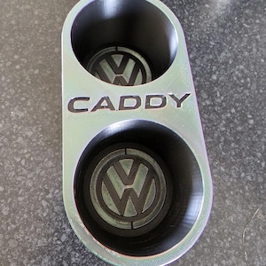 Puede incluir: Un portavasos negro y plateado con dos portavasos y la palabra "CADDY" impresa en la parte delantera. Cada portavasos tiene un logotipo de Volkswagen impreso en la parte inferior.
