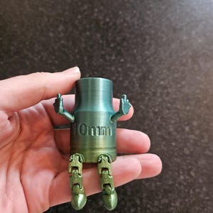 Puede incluir: Una figura verde y dorada impresa en 3D con piernas y brazos articulados, mostrando el dedo corazón. La figura es cilíndrica con el texto "10mm" impreso en el lateral.