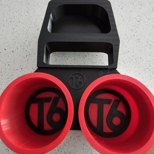 May include: A black and red 3D-printed organizer. The organizer has two red cup holders with a black "76" logo and a top compartment. The item is designed to hold items.