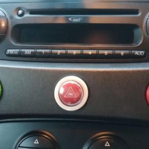 Puede incluir: Primer plano del salpicadero de un coche con reproductor de CD, controles de radio y varios botones. Los botones incluyen un botón "SPORT" verde, un botón de luces de emergencia rojo y un botón de faros rojos. El salpicadero es gris oscuro.