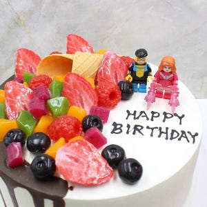 May include: A white frosted cake decorated with fresh strawberries, raspberries, blueberries, and colorful fruit jellies. Two Lego figures stand atop the cake, which is inscribed with the words "HAPPY BIRTHDAY".
