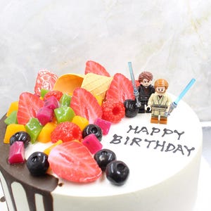 May include: A white frosted birthday cake decorated with fresh fruit, including strawberries, raspberries, and blueberries. The cake is topped with Lego figures and the words "HAPPY BIRTHDAY" in black.