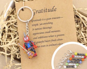 Könnte beinhalten: Ein Metallschlüsselanhänger mit einer roten und blauen Superhelden-Minifigur, die eine gelbe E-Gitarre hält, befestigt an einer Karte mit dem Text Gratitude und einem Gedicht. Ein kreisförmiger Einsatz zeigt sechs weitere Schlüsselanhänger mit Namensperlen.