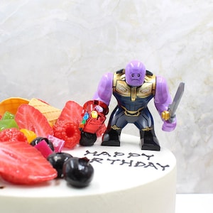 May include: A decorated cake with fresh fruit and a Thanos action figure. The cake is topped with strawberries, raspberries, and blueberries. The Thanos figure is purple, black, and gold, holding a sword and a red gauntlet. The words "HAPPY BIRTHDAY" are written on the cake.