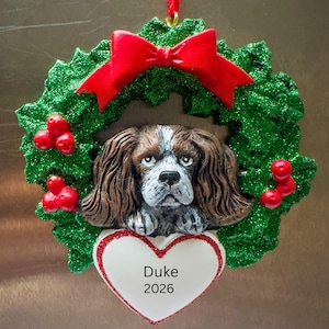 Puede incluir: Un adorno navideño con la cabeza de un perro Cavalier King Charles Spaniel marrón y blanco centrado en una corona de acebo verde brillante. Un lazo rojo corona la pieza y un corazón blanco con borde de purpurina roja muestra el texto Duke 2026.