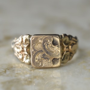 May include: A gold-colored ring with a rectangular face featuring an engraved floral design. The band has a textured, ornate pattern. The ring is set against a light-colored, marble-like surface.