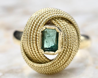 May include: A gold ring with a knotted design featuring a green emerald gemstone in the center.