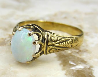 May include: A gold ring with an oval opal gemstone. The ring has an ornate, engraved band and a prong setting that holds the opal. The opal has a milky white color with flashes of iridescent green and blue.