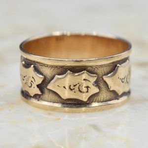 May include: A gold band ring with a decorative design of repeating stylized leaves. The ring is made of yellow gold and has a smooth, polished finish.