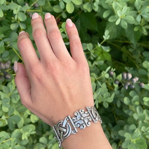 May include: A silver bracelet featuring a detailed, intricate design. The bracelet is made up of linked, interlocking pieces with a geometric pattern.