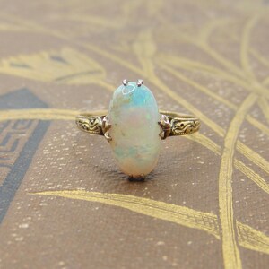 May include: A gold ring featuring an oval opal gemstone. The opal displays a mix of colors, including white, blue, and hints of pink. The ring's band has an ornate, detailed design. The opal measures approximately 0.75 inches in length.