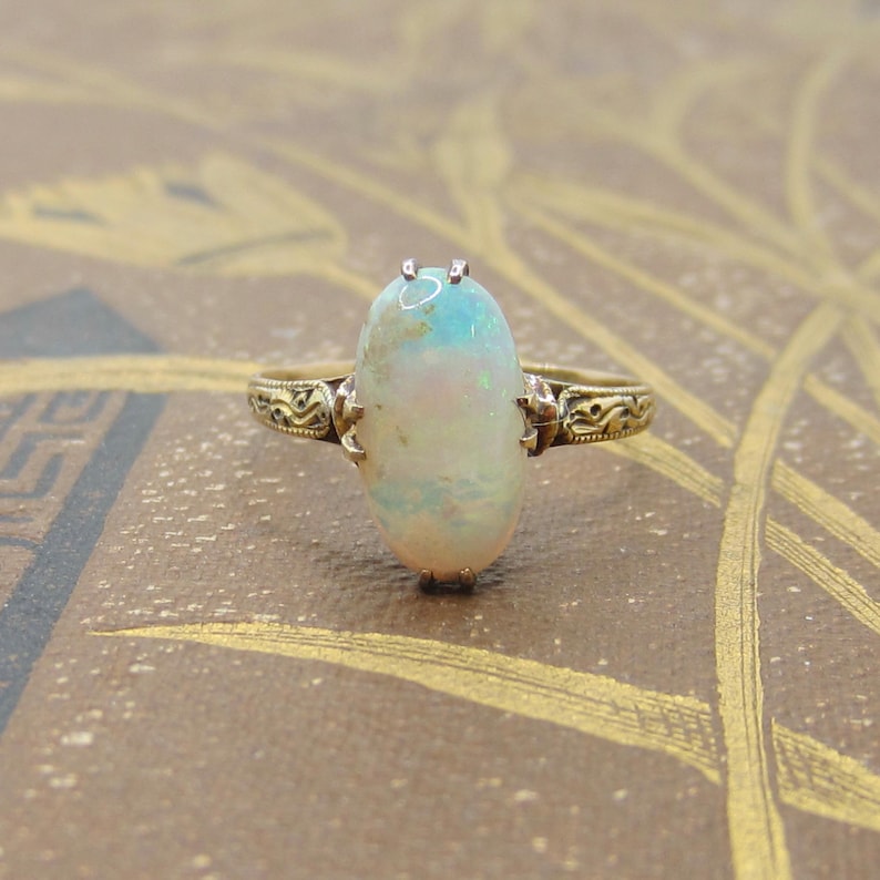 May include: A gold ring featuring an oval opal gemstone. The opal displays a mix of colors, including white, blue, and hints of pink. The ring's band has an ornate, detailed design. The opal measures approximately 0.75 inches in length.