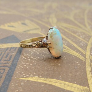 May include: A vintage gold ring featuring an oval opal gemstone. The opal has a milky white base with flashes of color. The ring's band and setting are detailed with intricate filigree patterns, showcasing a classic jewelry design.