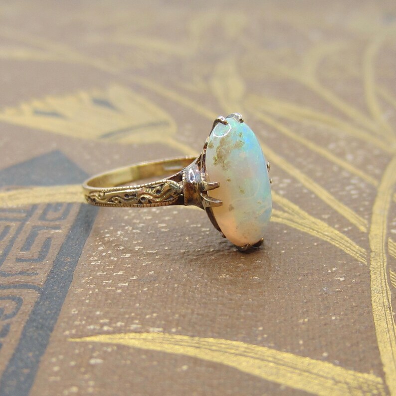 May include: A vintage gold ring featuring an oval opal gemstone. The opal has a milky white base with flashes of color. The ring's band and setting are detailed with intricate filigree patterns, showcasing a classic jewelry design.