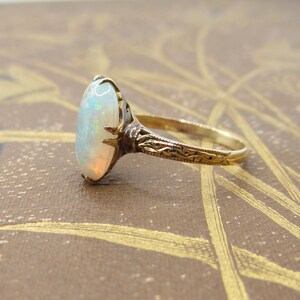 May include: A gold ring with an oval opal gemstone. The opal has a milky white base with flashes of color. The ring's band and setting are gold, with intricate engraved details. The ring is set against a brown background.