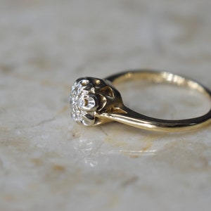 May include: A gold ring with a round, white diamond center stone surrounded by smaller diamonds. The ring has a delicate, intricate design.
