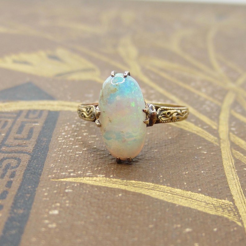 May include: A vintage gold ring featuring an oval opal gemstone. The opal displays a play of color with hints of blue, green, and pink. The ring has an ornate, detailed band with a filigree design, adding to its antique aesthetic.