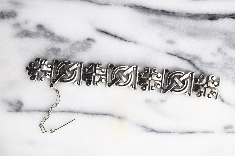May include: A silver bracelet with a detailed, intricate design. The bracelet is made of linked, interlocking pieces that form a repeating pattern.