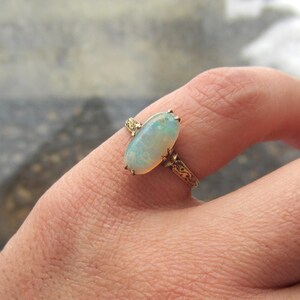 May include: A gold ring featuring an oval opal gemstone. The opal has a pale blue and white color. The ring's band and setting are gold-toned with intricate filigree detailing. The ring is shown on a finger.