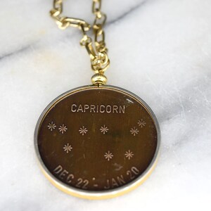 May include: A gold-toned Capricorn pendant with a matching chain. The pendant features the word "CAPRICORN" and the dates "DEC 22 - JAN 20" along with star constellations. The pendant is round and has a gold-colored border.