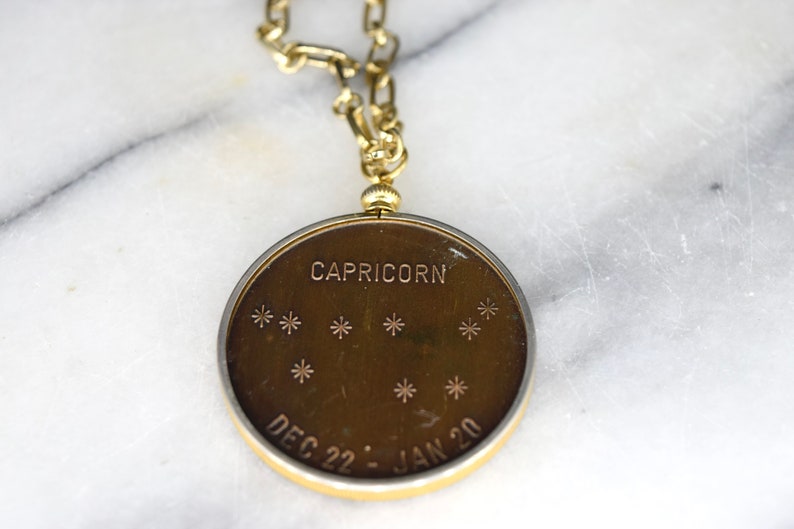 May include: A gold-toned Capricorn pendant with a matching chain. The pendant features the word "CAPRICORN" and the dates "DEC 22 - JAN 20" along with star constellations. The pendant is round and has a gold-colored border.